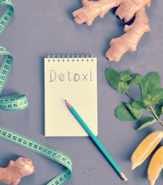 Gri arka plan üzerine defter ve kalem. Not defteri sayfasında yazıt Detoks! Dizüstü bilgisayarın yanında bir ölçüm bandı, zencefil kökü, limon ve nane var. Yukarıdan görüntüleyin. Close.