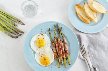 Bir tabak kızarmış yumurta ve pastırmalı kuşkonmaz. Tabağın yanında kızarmış baget var. Çerçevede su, taze kuşkonmaz ve çatal bıçak takımı ile bir bardak. Açık arka plan. Yukarıdan görüntüle.