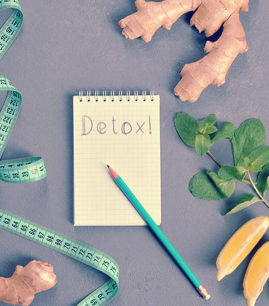 Gri arka plan üzerine defter ve kalem. Not defteri sayfasında yazıt Detoks! Dizüstü bilgisayarın yanında bir ölçüm bandı, zencefil kökü, limon ve nane var. Yukarıdan görüntüleyin. Close.