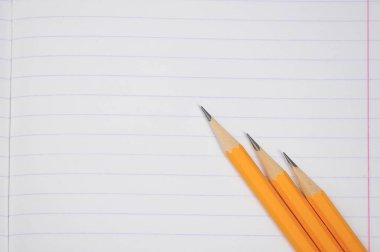 Not defteri sırada. Not defteri sayfası çerçevenin tüm alanını kaplar. Bir defter yaprağıüzerinde üç kalem. Yukarıdan görüntüleyin. Close.