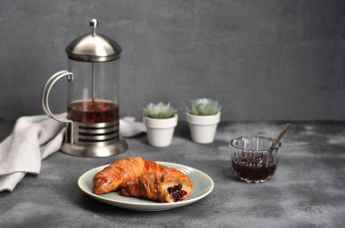 Tabakta iki çıtır kruvasan. Sonra bir fincan reçel ve çaydanlık çaydanlık. Çerçevede, bez peçete. Gri arka plan. Close.