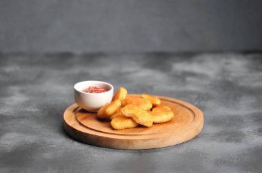 Yuvarlak bir tahta üzerinde gevrek kabuk ve patates kızartması ile tavuk fileto parçaları. Külçelerin yanında bir fincan ketçap var. Close. Gri arka plan.