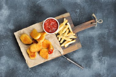 Tahta tahtada tavuk külçeleri ve patates kızartması. Bir sonraki bardak ketçap. Külçelerden biri çatala sabitlenmiş. Yukarıdan görüntüleyin. Close. Gri arka plan.