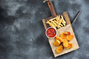 Tahta tahtada tavuk külçeleri ve patates kızartması. Bir sonraki bardak ketçap. Külçelerden biri çatala sabitlenmiş. Yukarıdan görüntüleyin. Gri arka plan. Metin için boş alan.