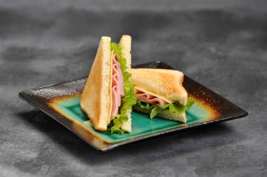 Bir tabakta üçgen şeklinde sandviçler. Sandviçin dolgusu jambon, peynir ve yapraklardan oluşur. Beyaz ekmek dilimleri kavrulur. Gri arka plan. Close.