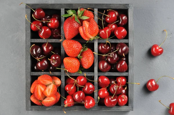 A box with ripe berries of strawberries and sweet cherries. Nearby a few berries of sweet cherry. View from above. Close-up. Gray background.