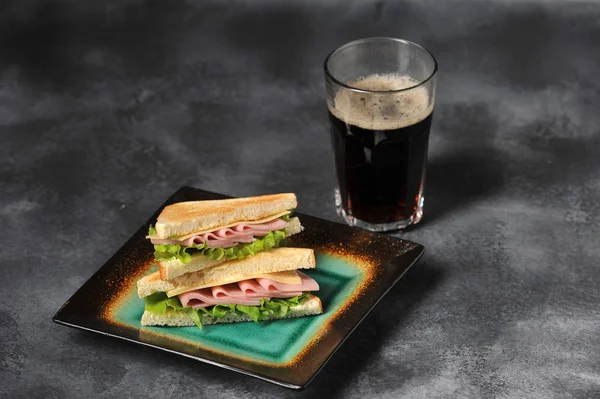 Sandwich with ham and cheese from white bread. Sliced bread is toasted. Near the glass with a carbonated drink. Gray background. Close-up.