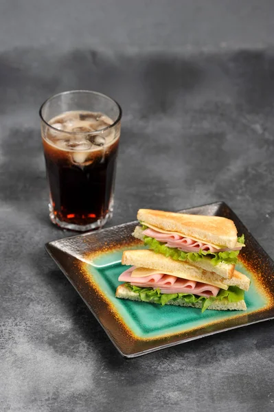 Beyaz ekmekten jambon ve peynirli sandviç. Dilimlenmiş ekmek kızartılır. Gazlı içecekle bardağın yanında. Gri arka plan. Close. Çerçevenin dikey yönü.