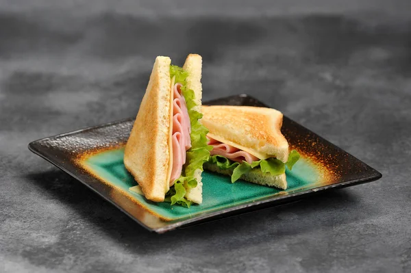 Bir tabakta üçgen şeklinde sandviçler. Sandviçin dolgusu jambon, peynir ve yapraklardan oluşur. Beyaz ekmek dilimleri kavrulur. Gri arka plan. Close.