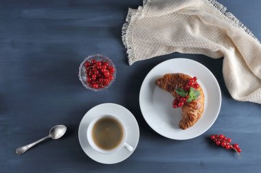 Hava kruvasan kırmızı frenk üzümü meyveleri ile dekore edilmiştir. Bir sonraki fincan sade kahve. Çerçevede, çilek, çay kaşığı, peçete ile bir vazo. Koyu ahşap arka plan. Yukarıdan görüntüleyin. Close.