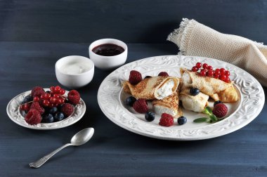 Krep, süzme peynirle doldurulmuş. Yemek ahududu, yaban mersini ve kırmızı kuş üzümü meyveleri ile dekore edilmiştir. Çerçeve reçel, ekşi krema, çilek ile bir tabak. Karanlık arka plan. Close.