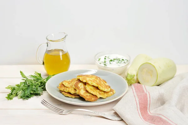 Small pancakes from a zucchini on a plate. Next to the dish there is a napkin, a fork, a bunch of parsley, a jug with olive oil and a cup with sour cream sauce. Light background. Close-up.
