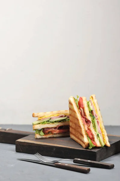 Classic club sandwich with ham and bacon on a wooden board. Next cup is a mixture of pickled miniature vegetables. Gray background. Close-up. Vertical orientation. Place under the text.