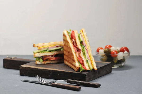 Classic club sandwich with ham and bacon on a wooden board. Next a cup of pickled vegetables. Gray background. Close-up.