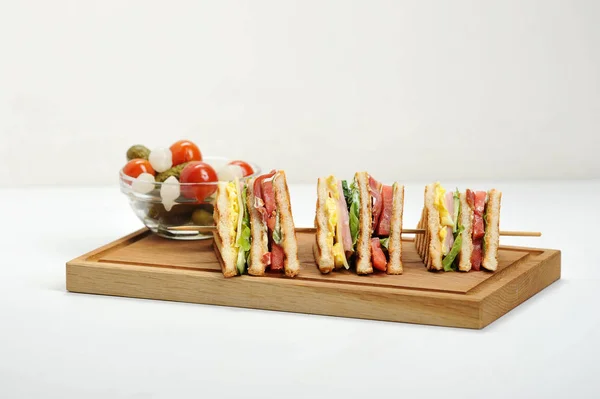 Sandwich on a light wooden board. The sandwich is cut into four pieces and strung on a wooden skewer. Next to a plate of pickled onions, tomato, cucumber. White background.