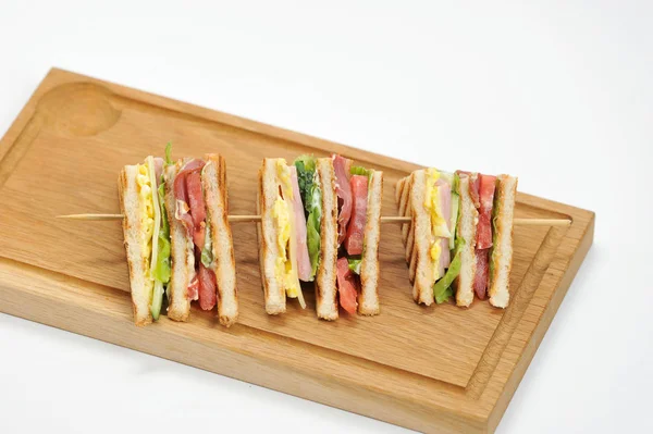 Sandwich on a light wooden board. In the filling of a sandwich ham, bacon, cheese, egg, tomato, cucumber, lettuce. The sandwich is cut into four pieces and strung on a wooden skewer. White background.