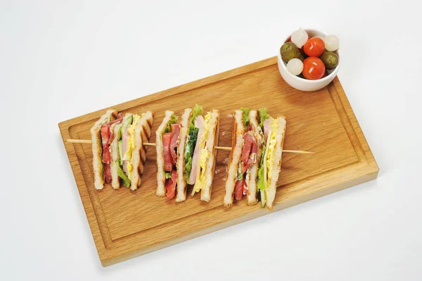 Hafif ahşap tahtada sandviç. Sandviç dört parçaya kesilir ve ahşap bir şiş üzerinde asılır. Bir tabak soğan, domates, salatalık tabağının yanında. Beyaz arka plan. Yukarıdan görüntüle.