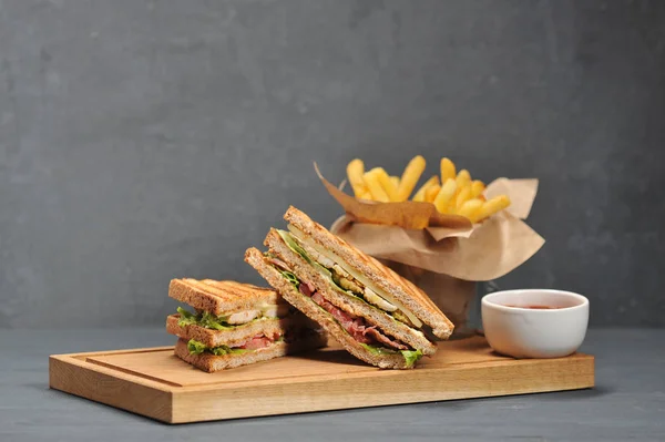 Club sandwich on a wooden board. Next to French fries and a cup of ketchup sauce. The filling of the sandwich consists of chicken breast, bacon, cheese, lettuce, tomato. Close-up. Gray background.