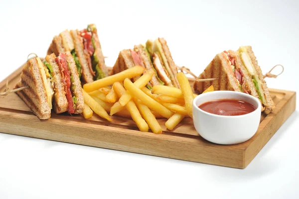 Club-sandwich and french fries on a light wooden board. Sandwiches are fastened with skewers. Next to the potatoes is a cup of ketchup. White background. Close-up.