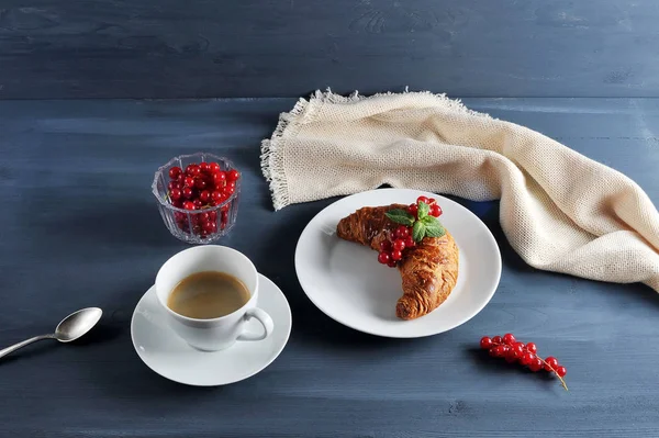 Hava kruvasan kırmızı frenk üzümü meyveleri ile dekore edilmiştir. Bir sonraki fincan sade kahve. Çerçevede, çilek, çay kaşığı, peçete ile bir vazo. Koyu ahşap arka plan. Close.