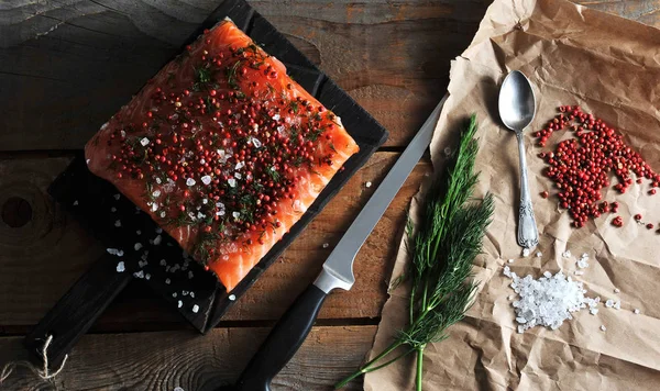 Karanlık tahtada bir parça taze somon. Balık deniz tuzu, pembe biber ve dereotu serpilir. Kraft kağıt tuz, karabiber, dereotu yanında. Çerçevede bir bıçak var. Ahşap arka plan. Yukarıdan görüntüle.