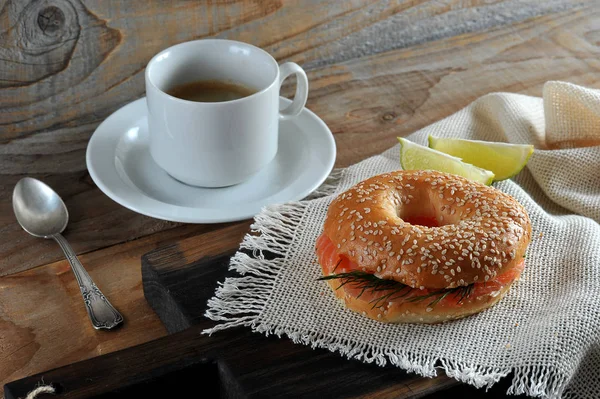 Beagle with salmon on a wooden board. Next two slices of lemon and a napkin. In the frame a cup with black coffee. Wooden background. Close-up. View from above.