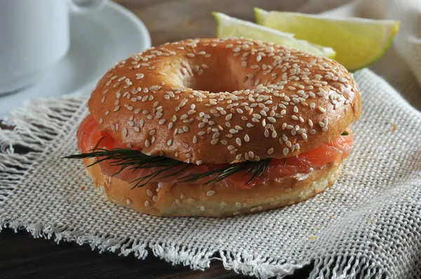 Beagle with salmon, soft cheese and dill on cloth napkin. There are two slices of lemon. In the frame a cup with black coffee. Close-up. Macro photography. View from above.