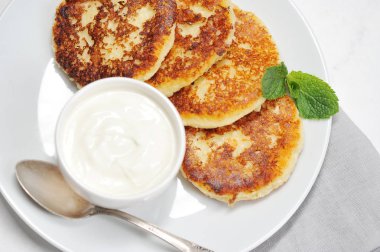 Peynirli kekler beyaz tabakta. Peynirli keklerin yanında bir fincan ekşi krema ve bir kaşık var. Peynirli kekler taze nane yaprağıyla süslenir. Yukarıdan bak. Yakın plan. Makro fotoğrafçılık.