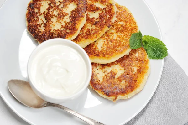 Peynirli kekler beyaz tabakta. Peynirli keklerin yanında bir fincan ekşi krema ve bir kaşık var. Peynirli kekler taze nane yaprağıyla süslenir. Yukarıdan bak. Yakın plan. Makro fotoğrafçılık.