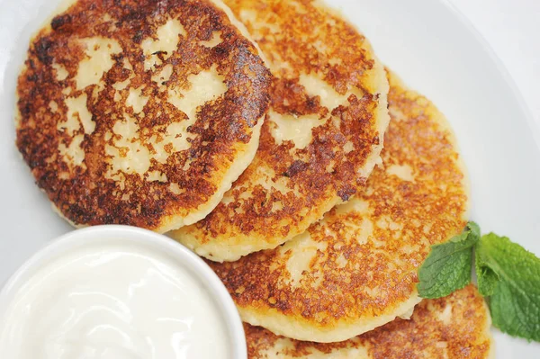 Beyaz tabakta cheesecakes. Cheesecakes yanında ekşi krema bir fincan. Syrniki taze nane yaprağı ile dekore edilmiştir. Yukarıdan görüntüleyin. Close. Makro fotoğrafçılık.