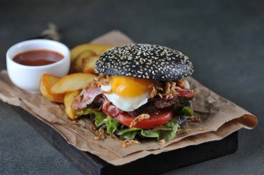 Kızarmış yumurtalı siyah burger. Pastırma ve soğan cipsi dolduran hamburgerde. Hamburgerin yanında rustik bir patates. Karanlık arka plan. Close. Close.
