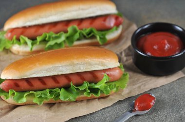 Zanaat kağıt üzerinde iki sosisli sandviç. Ketçap soslu bir fincan ve soslu bir kaşık. Close. Koyu arka plan.