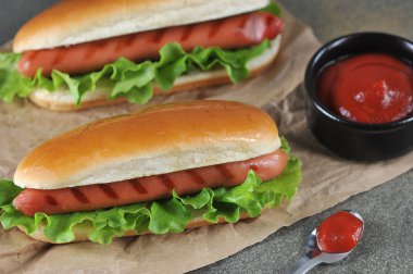 Zanaat kağıt üzerinde iki sosisli sandviç. Ketçap soslu bir fincan ve soslu bir kaşık. Close. Koyu arka plan.