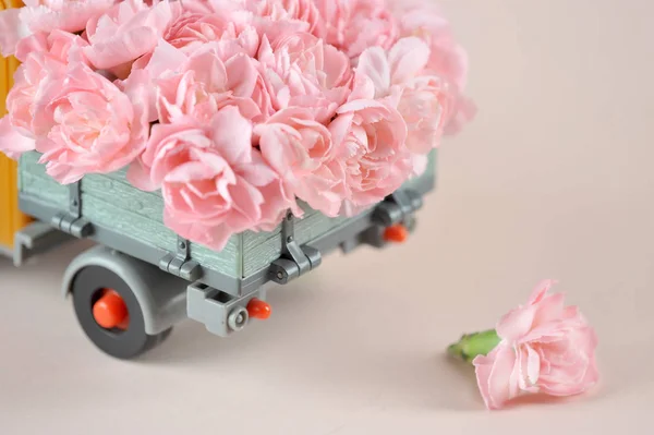 Toy car with pink flowers in the back. Light pink background. Close-up. Place for text. The concept of the original congratulations on the holiday.