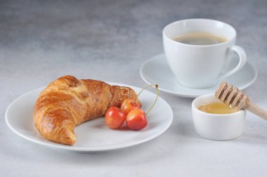 Beyaz tabakta kruvasan ve sarı tatlı kiraz meyveleri. Bir fincan sade kahve ve bal bileşimi tamamlar. Lezzetli klasik kahvaltı konsepti. Close. Açık arka plan.