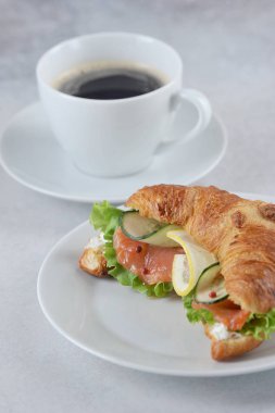 Somon sandviç ve bir fincan kahve gibi kruvasan. Sandviç dolgusuna yumuşak peynir, tuzlu somon, salata, salatalık ve limon dilimleri dahildir. Açık arka plan. Close. Dikey yönlendirme.