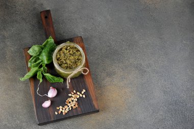 Cam bir kavanozda pesto yeşil sos. Sarımsak, fesleğen yeşillikleri ve çam fıstığı bir kavanoz yanında ahşap bir tahta üzerinde. Close. Gri arka plan. Yukarıdan görüntüleyin. Metin için boş alan.