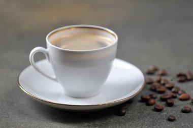 Koyu bir yüzeyde beyaz bir fincan kahve. İnce porselen fincan ve tabak. Masanın yüzeyinde kahve çekirdekleri. Close.