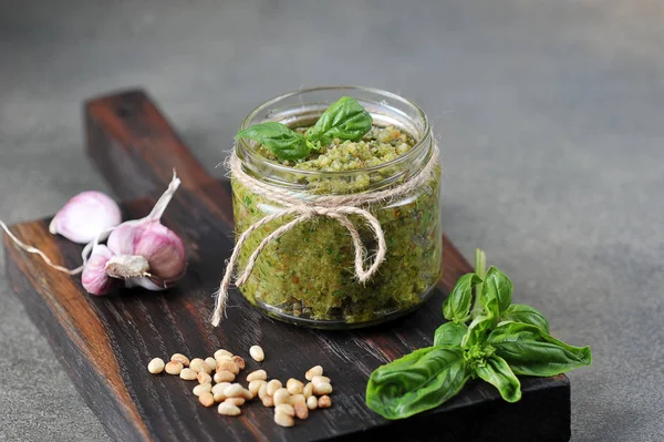 Pesto green sauce in a glass jar. On a wooden board next to a jar of garlic, basil greens and pine nuts. Close-up. Gray background.