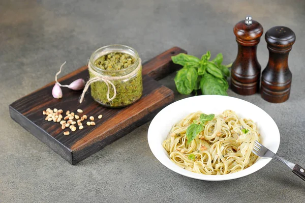 Pasta with pesto and basil. Nearby a jar of pesto, garlic, nuts and seasonings. Lunch table setting concept. Dark background. Close-up.