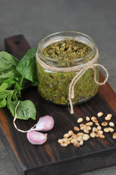 Pesto green sauce in a glass jar. On a wooden board next to a jar of garlic, basil greens and pine nuts. Close-up. Gray background. The vertical orientation of the frame.