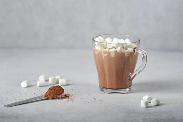 Cocoa with marshmallows in a transparent glass. Marshmallows are scattered on a light surface and a spoonful of cocoa powder lies. Close-up.