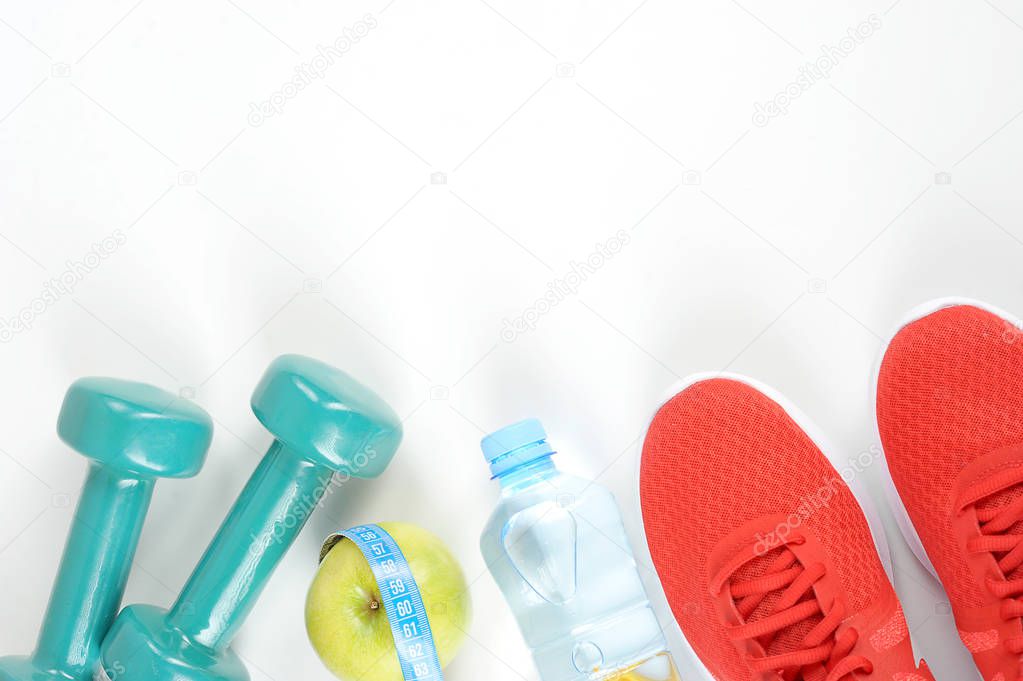 Dumbbells, green apple, drinking water, a measuring tape and sneakers on a white background. View from above. Free space for text.