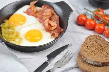 Tavada pastırma ile kızarmış yumurta. Koyu ekmek dilimleri, çatal bıçak takımı, çeri domates ve baharatlar kompozisyonu tamamlar. Açık arka plan. Close. Makro çekimi.