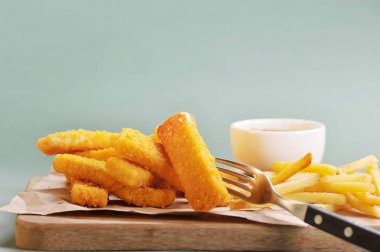 Hafif tahta bir tahtada balık çubukları ve patates kızartması. Sos ek olarak kullanılır. Balık çubuğu çatalla delinmiş. Açık arka plan. Close. 