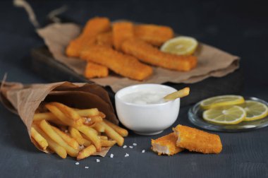 Koyu ahşap bir arka plan üzerinde balık çubukları ve patates kızartması. Kraft kağıdında patates kızartması. Sos ve limon dilimleri ek olarak kullanılır. Close. 