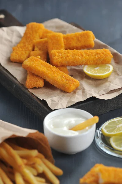 Fish sticks and french fries on a dark wooden background. French fries in a packet of Kraft paper. Sauce and lemon slices are used as a supplement. Close-up. Vertical frame orientation.