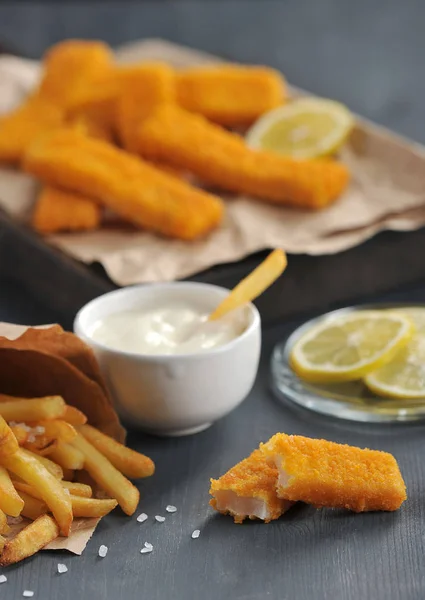 Fish sticks and french fries on a dark wooden background. French fries in a packet of Kraft paper. Sauce and lemon slices are used as a supplement. Close-up. Vertical frame orientation. Macro shooting