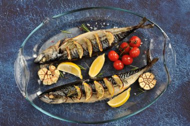 Uskumru ızgarada pişirilir. Tabakta iki balık var. Hazırlanmasında biberiye, limon, sarımsak, kiraz domates kullanılır. Yukarıdan görüntüleyin. Close. Koyu arka plan.