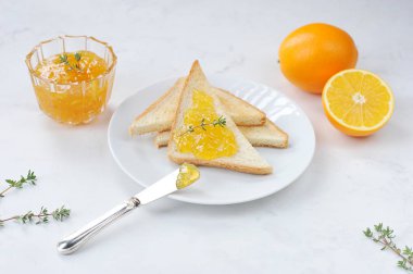 Beyaz bir tabakta portakal reçeli ile tost. Tabağın yanında bir kase portakal reçeli, iki portakal ve çatal bıçak takımı var. Açık arka plan. Close.